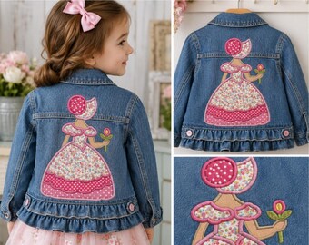 Può includere: Una giacca in denim blu con una grande applicazione sul retro raffigurante una figura con un abito floreale e una cuffietta rosa a pois, che tiene in mano un fiore. La giacca presenta un orlo arricciato, un colletto classico e bottoni rosa.