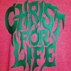 以下が含まれることがあります： ピンクのシャツに緑色の文字で「Christ for Life」とスタイリッシュなフォントで書かれています。