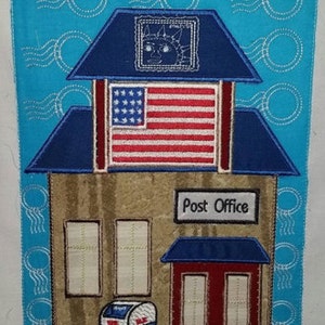 Puede incluir: Aplique bordado de un edificio de correos con techo azul, puerta roja y dos ventanas. El edificio tiene un letrero que dice "Post Office" y una bandera estadounidense sobre la puerta. Hay un buzón azul con las palabras "US Mail" en frente del edificio.
