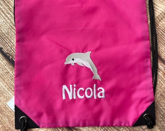 Op de afbeelding: Roze trekkoordtas met een geborduurde grijze dolfijn en de naam "Nicola" in wit.