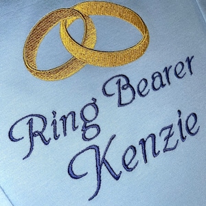 Op de afbeelding: Een lichtblauwe baby romper met gouden geborduurde trouwringen en de tekst "Ring Bearer Kenzie" in blauw cursief.