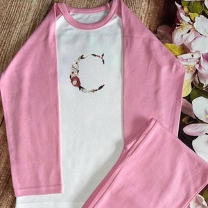 May include: Pink and white long-sleeved pajamas with a floral embroidered letter C on the front.