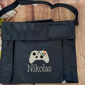 May include: A dark blue canvas bag with a white embroidered video game controller and the name "Nikolas" underneath. The bag has a flap with a small pocket on the front.