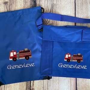 以下が含まれることがあります： 赤い消防車と「Genevieve」という名前が刺繍された青いバッグが2つ。1つは巾着式のバックパック、もう1つは大きめのトートバッグです。