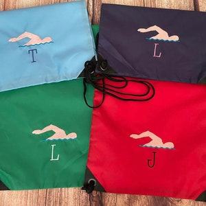 May include: Four drawstring bags in different colours with embroidered swimmer designs. The bags are blue, green, red, and navy blue. The bags have the letters T, L, J, and L embroidered on them.