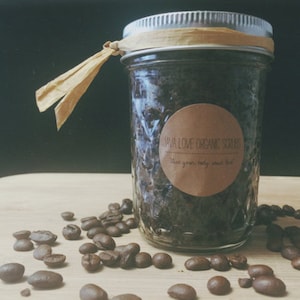Pode incluir: Um frasco de vidro cheio de um esfoliante de café marrom escuro. O frasco tem um rótulo marrom que diz "Java Love Organic Scrubs" e "Give your body some love". O frasco está sentado em uma superfície de madeira com grãos de café espalhados ao redor.
