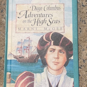 Op de afbeelding: Een boek getiteld "Diego Columbus Adventures on the High Seas" door Marni McGee. De omslag toont een illustratie van een jonge jongen die een rood-witte hoed en een rood-wit shirt draagt, staand op een houten pier. Een schip met een wit zeil en een rood kruis is op de achtergrond te zien.