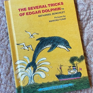 May include: A yellow book cover with a cartoon illustration of a dolphin jumping out of the water. The dolphin is black and gray and is jumping over a small boat. The book title is "The Several Tricks of Edgar Dolphin" by Nathaniel Benchley. The book is an "I Can Read Book".