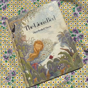 May include: A hardcover children's book titled The Lion's Bed by Diane Redfield Massie. The cover features a colorful illustration of a lion resting in a jungle setting with various plants, a bird, and an elephant, rendered in a soft, painterly style.