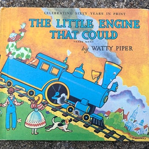Könnte beinhalten: Ein Vintage-Kinderbuchcover mit einer blauen Lokomotive mit einem lächelnden Gesicht, die einen Wagen zieht. Der Buchtitel lautet "The Little Engine That Could" von Watty Piper. Das Cover zeigt außerdem einen Clown in einem grün-weißen Kostüm, ein Mädchen in einem rot-weißen Kleid und einen Hund.