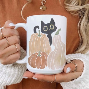 Puede incluir: Una taza de cer&aacute;mica blanca con un dise&ntilde;o de gato negro y calabazas. El gato mira hacia la derecha y tiene ojos grandes y redondos. Las calabazas son de color naranja y marr&oacute;n y tienen un tema oto&ntilde;al. La taza tiene un asa negra.