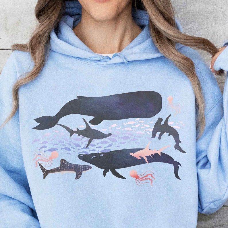 K&ouml;nnte beinhalten: Ein hellblauer Hoodie mit einem grafischen Druck verschiedener Meerestiere, darunter ein Wal, ein Hai, ein Hammerhai und ein Rochen. Die Grafik ist in Blaut&ouml;nen und Schwarz gehalten, mit rosa Akzenten f&uuml;r die Quallen.