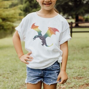 K&ouml;nnte beinhalten: Ein wei&szlig;es T-Shirt mit einem Regenbogen-Drachen-Motiv. Der Drache fliegt mit ausgebreiteten Fl&uuml;geln. Das Shirt ist aus einem weichen, bequemen Stoff.