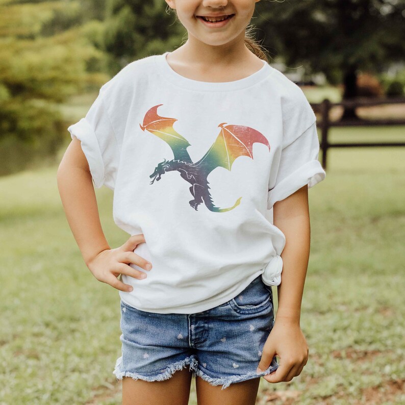K&ouml;nnte beinhalten: Ein wei&szlig;es T-Shirt mit einem Regenbogen-Drachen-Motiv. Der Drache fliegt mit ausgebreiteten Fl&uuml;geln. Das Shirt ist aus einem weichen, bequemen Stoff.