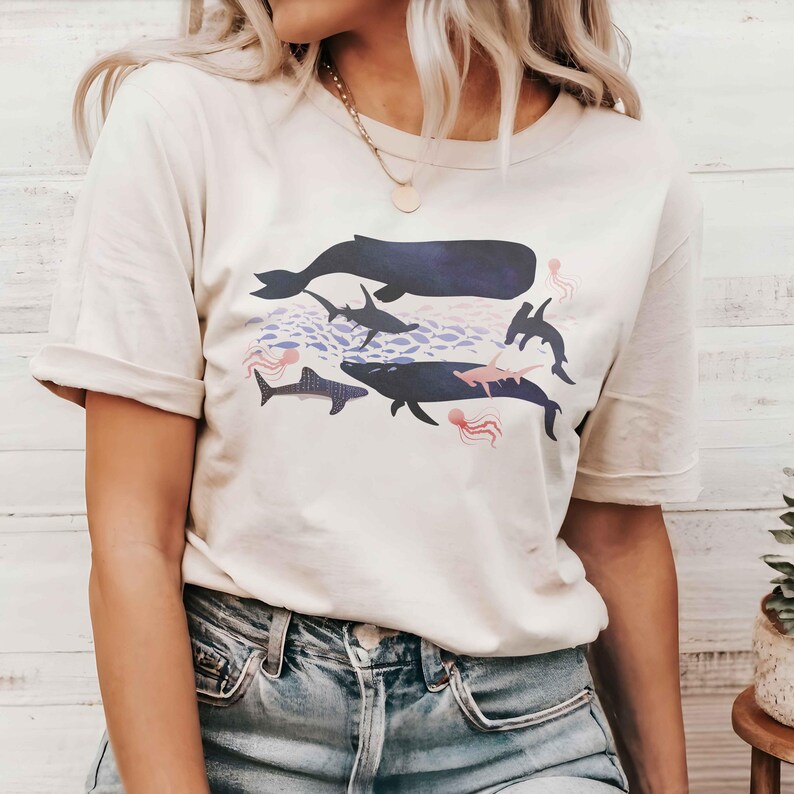 K&ouml;nnte beinhalten: Ein wei&szlig;es T-Shirt mit einem grafischen Aufdruck verschiedener Meerestiere, darunter ein Wal, ein Hai und Quallen. Die Grafik ist in Blaut&ouml;nen, Rosa und Lila gehalten.