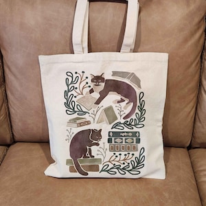 Puede incluir: Una bolsa de lona blanca con un dise&ntilde;o de dos gatos negros leyendo libros. El dise&ntilde;o incluye hojas verdes, libros marrones y una pila de libros con un patr&oacute;n de rayas marrones y blancas.