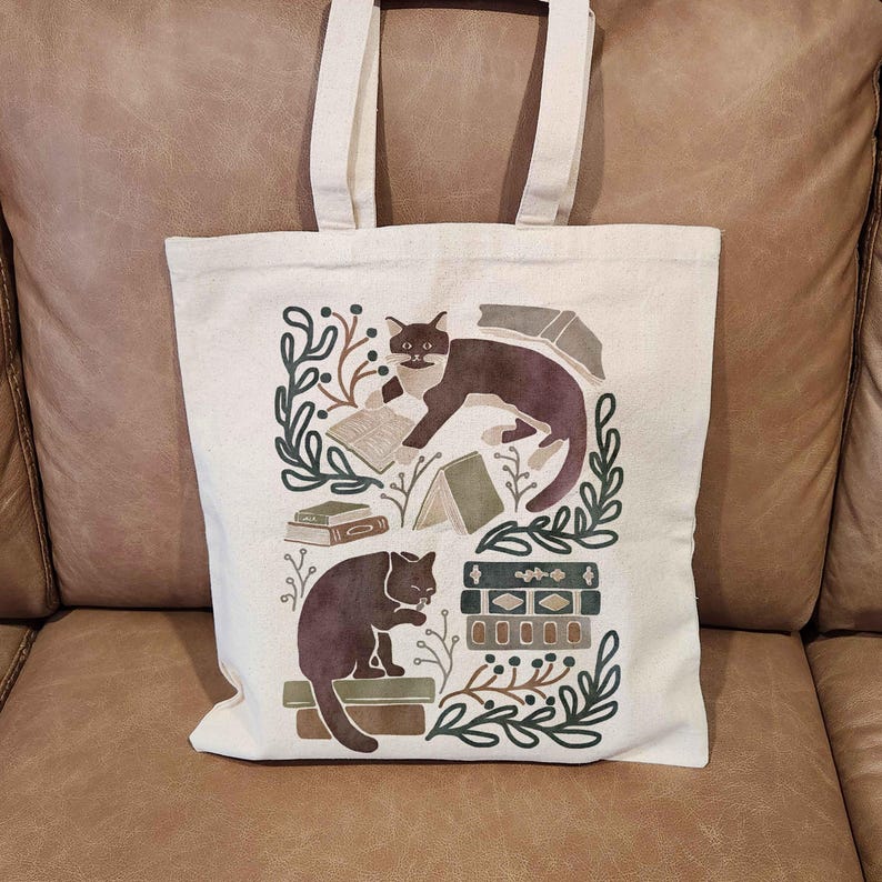 Puede incluir: Una bolsa de lona blanca con un dise&ntilde;o de dos gatos negros leyendo libros. El dise&ntilde;o incluye hojas verdes, libros marrones y una pila de libros con un patr&oacute;n de rayas marrones y blancas.