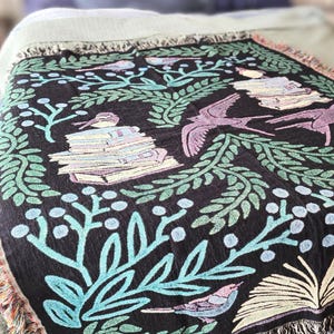 A woven blanket featuring a whimsical dark fantasy design of birds, books, and botanical elements in shades of green, purple, and blue. Available with a white or black background, the intricate illustration resembles a storybook or enchanted library scene, perfect for book lovers. The high-quality woven fabric adds texture and warmth, making it ideal as a cozy throw, reading nook accent, or wall tapestry