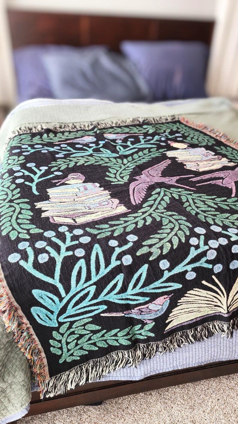 A woven blanket featuring a whimsical dark fantasy design of birds, books, and botanical elements in shades of green, purple, and blue. Available with a white or black background, the intricate illustration resembles a storybook or enchanted library scene, perfect for book lovers. The high-quality woven fabric adds texture and warmth, making it ideal as a cozy throw, reading nook accent, or wall tapestry