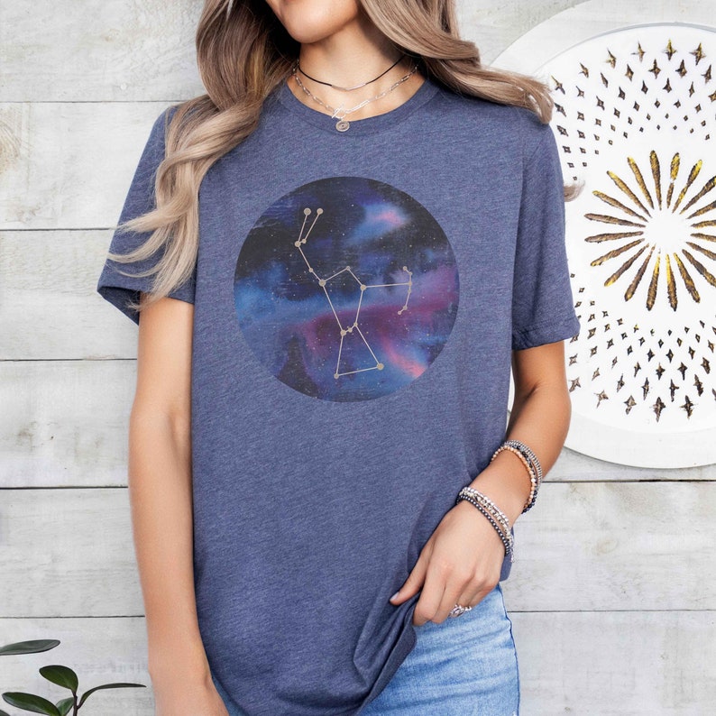 K&ouml;nnte beinhalten: Ein marineblaues T-Shirt mit einem Galaxie-Druck und einem goldenen Sternbild-Design. Das Sternbild hat die Form eines B&auml;ren.