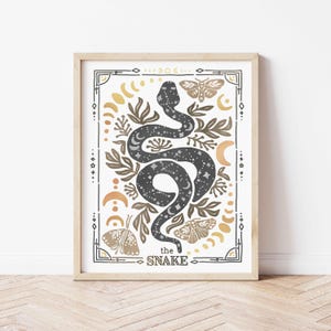 K&ouml;nnte beinhalten: Ein gerahmter Druck mit einer schwarzen Schlange mit wei&szlig;en Sternen, umgeben von goldenen und braunen floralen Designs, Monden und Schmetterlingen. Der Text "the SNAKE" befindet sich am unteren Rand des Drucks.