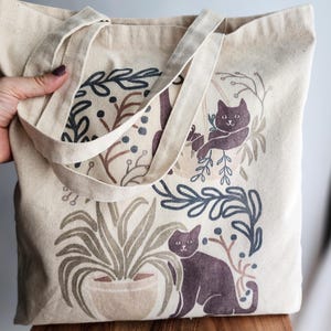 Whimsical canvas tote bag featuring illustrated cats lounging among potted houseplants. Soft watercolor tones in green and beige add a cozy, botanical feel. Perfect for cat lovers and plant enthusiasts