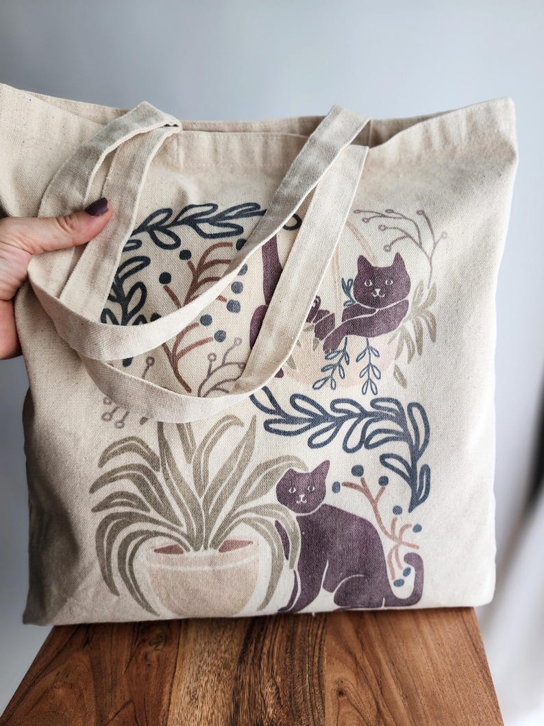 Whimsical canvas tote bag featuring illustrated cats lounging among potted houseplants. Soft watercolor tones in green and beige add a cozy, botanical feel. Perfect for cat lovers and plant enthusiasts