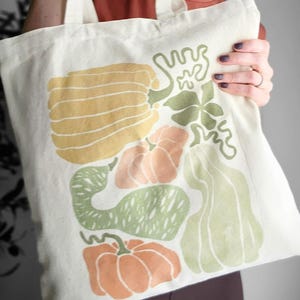 Puede incluir: Bolso tote de color crema con un estampado colorido de varias calabazas y calabazas en tonos amarillos, naranjas y verdes. El dise&ntilde;o incluye una calabaza grande y rayada, varias calabazas y otras formas de calabaza. El bolso tiene dos asas.