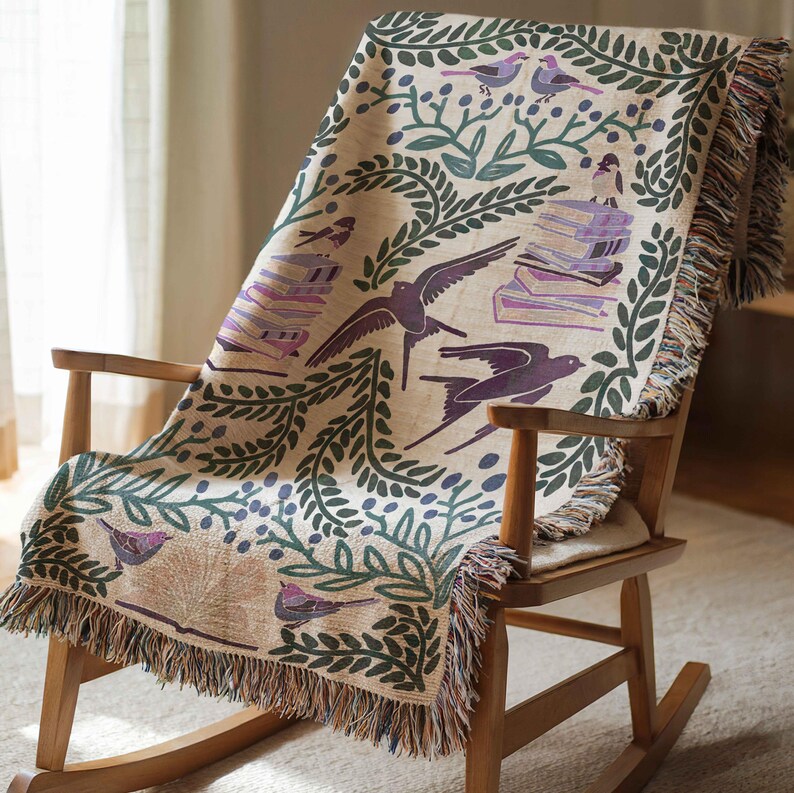 May include: A woven throw blanket with a cream background featuring a repeating pattern of green leaves, purple birds, and stacks of purple books. The blanket is draped over a wooden rocking chair.