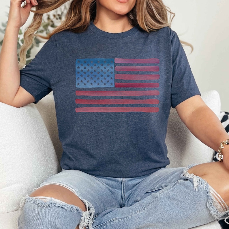 May include: A navy blue t-shirt with a watercolor-style American flag design. The flag is in shades of red, white, and blue.
