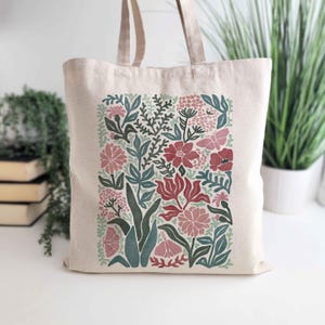 K&ouml;nnte beinhalten: Beige Canvas-Tragetasche mit floralem Design. Das Design umfasst rote, rosa und gr&uuml;ne Blumen und Bl&auml;tter. Die Tragetasche hat zwei beige Griffe und eignet sich zum Tragen von Lebensmitteln oder anderen Gegenst&auml;nden.
