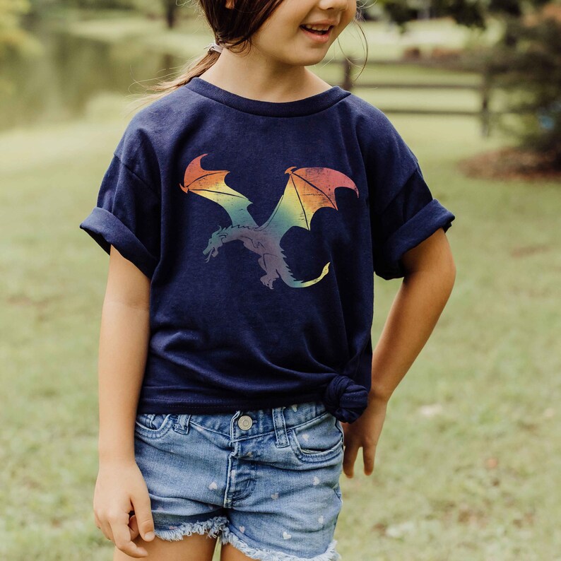 K&ouml;nnte beinhalten: Ein junges Kind tr&auml;gt ein marineblaues T-Shirt mit einem Regenbogen-Drachen-Motiv. Der Drache fliegt mit ausgebreiteten Fl&uuml;geln. Das Kind tr&auml;gt auch Jeans-Shorts.