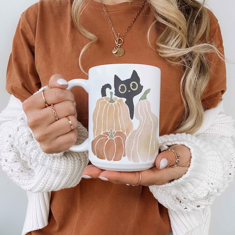 Puede incluir: Una taza de cer&aacute;mica blanca con un dise&ntilde;o de gato negro y calabazas. El gato mira hacia la derecha y tiene ojos grandes y redondos. Las calabazas son de color naranja y marr&oacute;n y tienen un tema oto&ntilde;al. La taza tiene un asa negra.