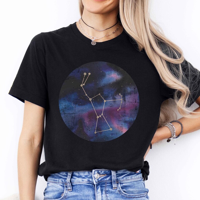 K&ouml;nnte beinhalten: Ein schwarzes T-Shirt mit einem goldenen Konstellationsdesign auf einem Aquarell-Galaxie-Hintergrund. Die Konstellation hat die Form des Gro&szlig;en B&auml;ren, auch bekannt als Gro&szlig;er Wagen.