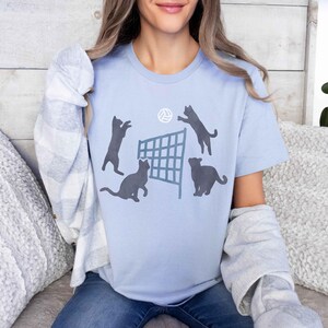 May include: A light blue t-shirt with a graphic of four cats playing volleyball. The cats are black silhouettes and the volleyball net is gray. The volleyball is white.