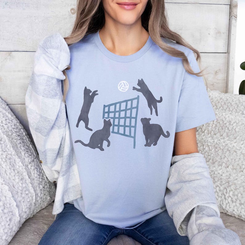 May include: A light blue t-shirt with a graphic of four cats playing volleyball. The cats are black silhouettes and the volleyball net is gray. The volleyball is white.