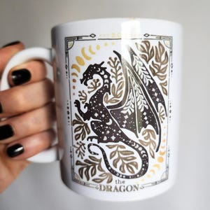 Watercolor celestial dragon mug featuring a mystical dragon surrounded by stars, moons, and botanical elements, framed with an elegant tarot-style border — fantasy-inspired boho coffee cup design.