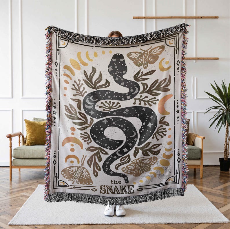 K&ouml;nnte beinhalten: Eine gewebte Tapisserie-Decke mit cremefarbenem Hintergrund und einem schwarzen Schlangenmuster. Die Schlange ist von goldenen und braunen floralen Elementen, Monden und Schmetterlingen umgeben. Der Text "the snake" ist unten auf der Decke aufgedruckt.