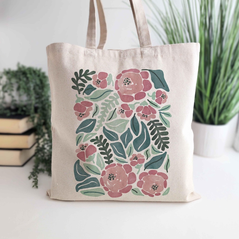 K&ouml;nnte beinhalten: Beige Canvas-Tragetasche mit floralem Design. Das Design umfasst rosa Rosen, gr&uuml;ne Bl&auml;tter und kleinere rosa Bl&uuml;ten. Die Tragetasche hat zwei beige Griffe und eignet sich zum Tragen verschiedener Gegenst&auml;nde.