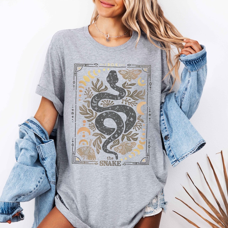 May include: Gray t-shirt with a graphic print of a black snake with stars and moons. The graphic is surrounded by a gold frame and the text "the snake" is below the graphic.