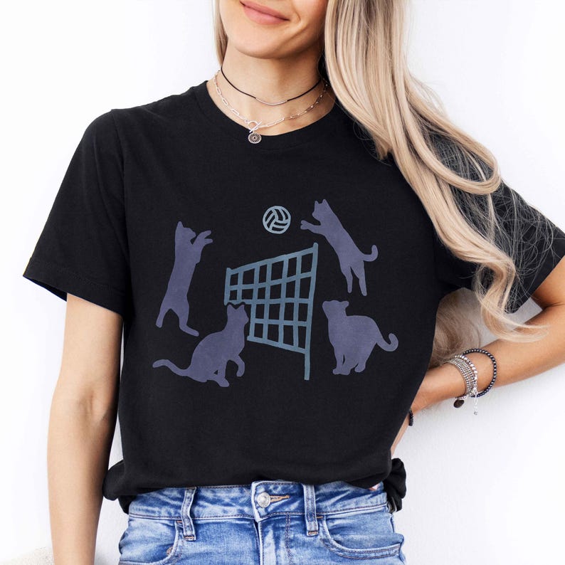 May include: Black t-shirt with a graphic of four cats playing volleyball. The cats are silhouetted in black and the volleyball net is blue.