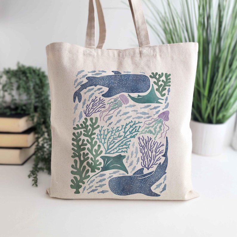 May include: A white canvas tote bag with a colorful illustration of a whale, fish, and coral reef. The design is in shades of blue, green, and purple.