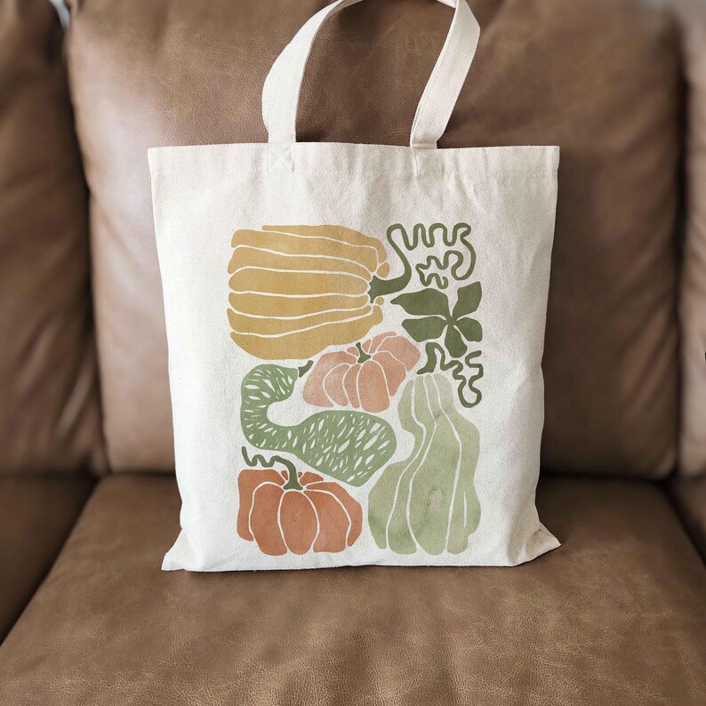 Puede incluir: Bolso tote de color crema con una colorida ilustraci&oacute;n de varias calabazas y calabacines en tonos naranja, verde y amarillo. El bolso tiene dos asas blancas y est&aacute; hecho de un material de lona resistente.