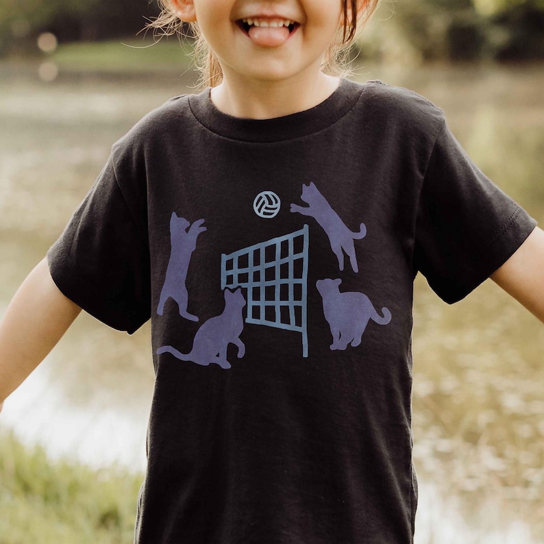 May include: A black t-shirt with a graphic of cats playing volleyball. The cats are jumping and reaching for a volleyball over a net. The graphic is in shades of blue.