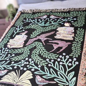 A woven blanket featuring a whimsical dark fantasy design of birds, books, and botanical elements in shades of green, purple, and blue. Available with a white or black background, the intricate illustration resembles a storybook or enchanted library scene, perfect for book lovers. The high-quality woven fabric adds texture and warmth, making it ideal as a cozy throw, reading nook accent, or wall tapestry