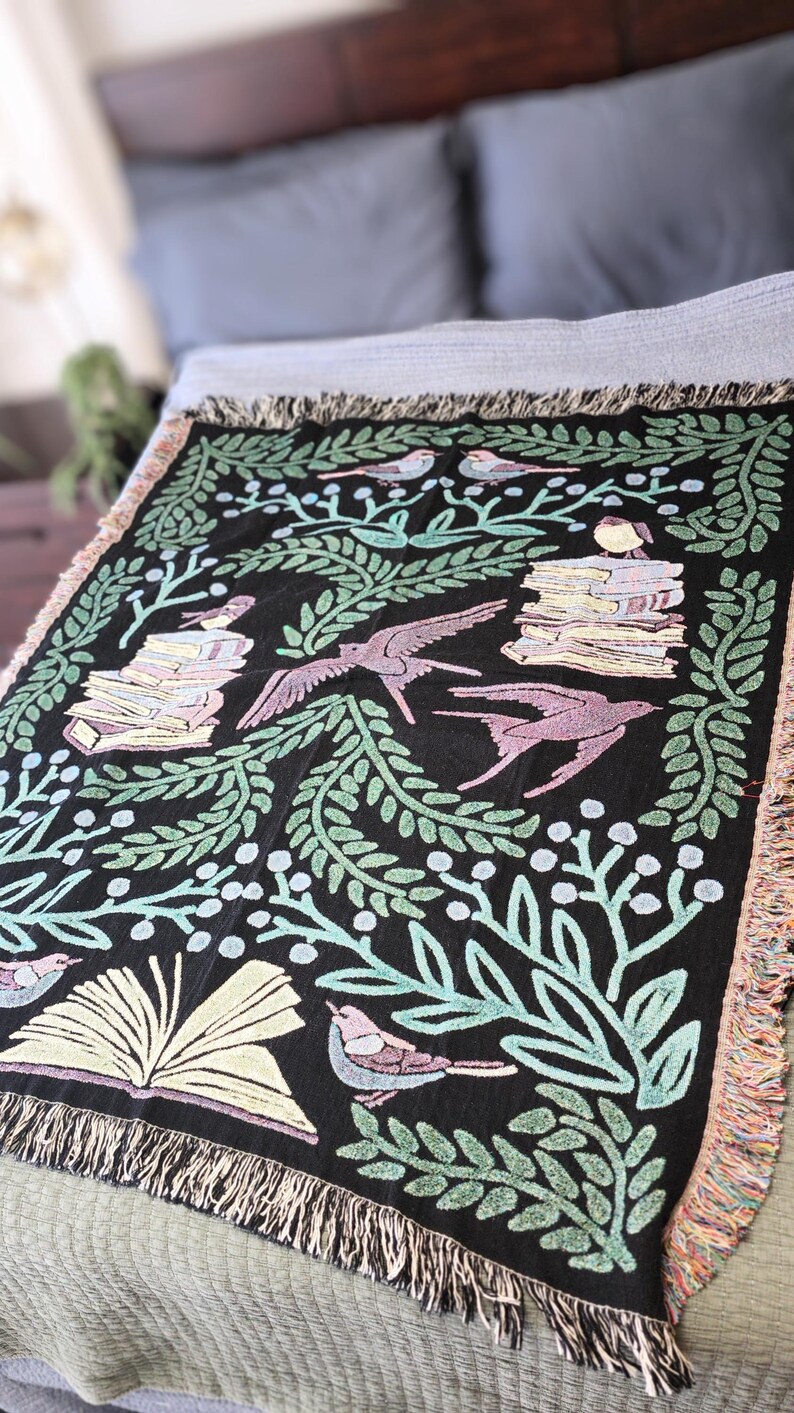A woven blanket featuring a whimsical dark fantasy design of birds, books, and botanical elements in shades of green, purple, and blue. Available with a white or black background, the intricate illustration resembles a storybook or enchanted library scene, perfect for book lovers. The high-quality woven fabric adds texture and warmth, making it ideal as a cozy throw, reading nook accent, or wall tapestry