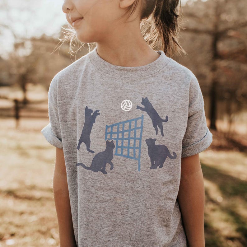 May include: A gray t-shirt with a graphic of four cats playing volleyball. The cats are black silhouettes and the volleyball net is blue. The volleyball is white.