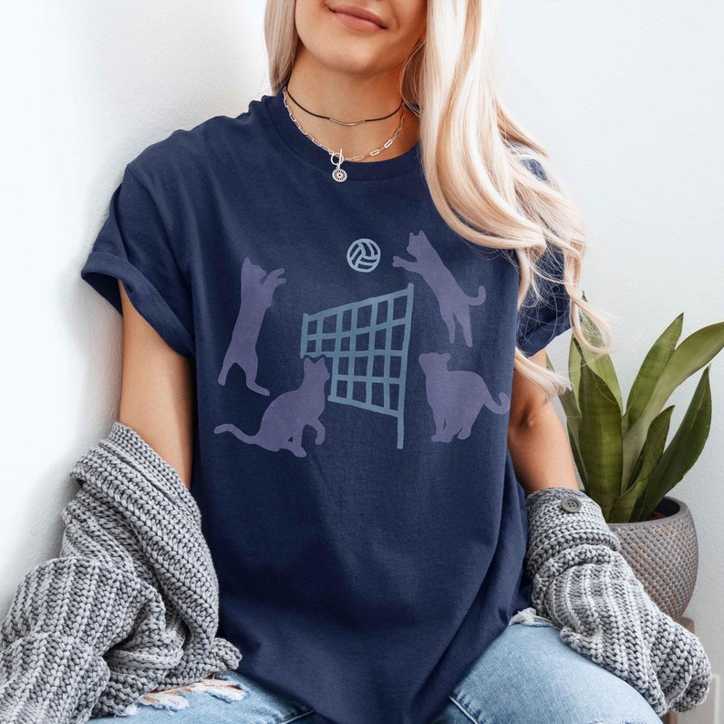May include: A navy blue t-shirt with a graphic of cats playing volleyball. The cats are silhouetted in a light blue color. The volleyball net is also light blue. The volleyball is a light blue circle with a white star in the center.