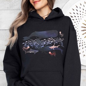 K&ouml;nnte beinhalten: Schwarzer Hoodie mit einem bunten Grafikdruck verschiedener Meerestiere, darunter ein Wal, Haie, Quallen und Fische. Die Grafik ist ein mehrschichtiges Design mit einem dunkelblauen Hintergrund.