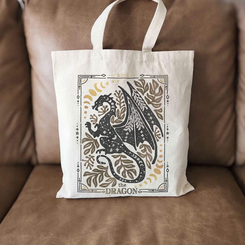 Puede incluir: Una bolsa de tela blanca con una silueta negra de un drag&oacute;n rodeada de dise&ntilde;os florales y lunares. El texto "the DRAGON" est&aacute; impreso debajo del drag&oacute;n.